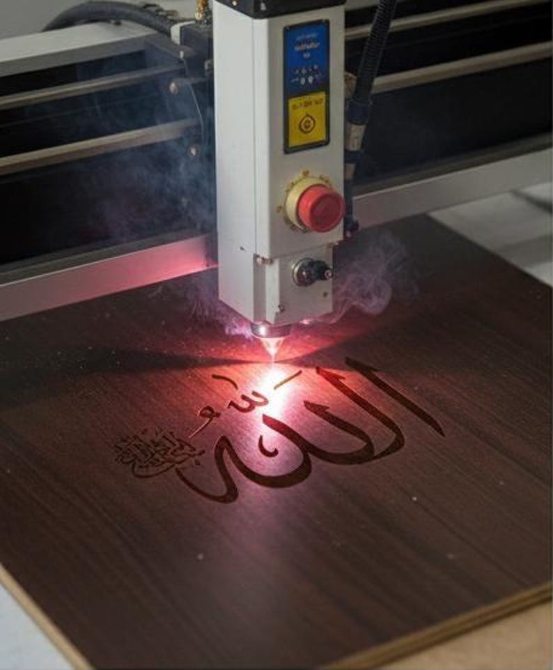K&ouml;nnte beinhalten: Eine Lasergravurmaschine graviert arabische Kalligraphie auf eine dunkle Holzoberfl&auml;che. Die wei&szlig;en und grauen Komponenten der Maschine sind sichtbar, mit einem roten Not-Aus-Knopf. Der Gravurprozess erzeugt Rauch und ein hellrotes Licht.
