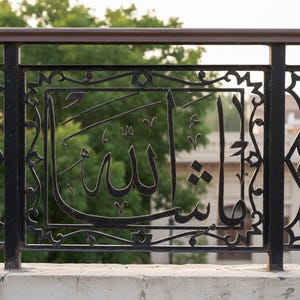 K&ouml;nnte beinhalten: Schwarzes schmiedeeisernes Gel&auml;nder mit arabischer Kalligraphie. Das Design zeigt verzierte geometrische Muster und das Wort "Allah" in eleganter Schrift. Der Hintergrund zeigt gr&uuml;nes Laub und ein Geb&auml;ude.
