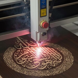 K&ouml;nnte beinhalten: Eine Lasergravurmaschine graviert auf einer dunklen Holzoberfl&auml;che aufwendige arabische Kalligraphie. Der Laser emittiert einen hellroten Strahl, der Rauch erzeugt. Das gravierte Design hat einen helleren Braunton.