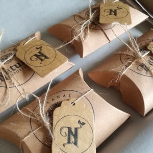 Peut inclure: Quatre boîtes cadeaux en papier brun avec de la ficelle et une étiquette en papier brun avec un N noir et un petit motif de feuille. L'étiquette indique "Naturel" en bas.