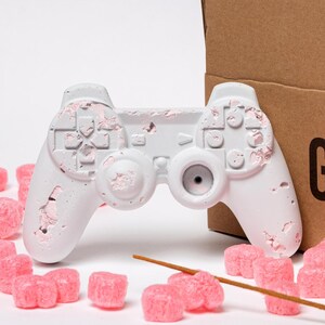 Op de afbeelding: Een witte wierookhouder in de vorm van een gamecontroller, met roze accenten, naast een bruine kartonnen doos. De doos is voorzien van het opschrift "GYPSTER INCENSE HOLDER". Roze verpakkingschips en een wierookstokje liggen verspreid.