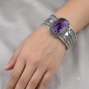 Peut inclure: Un bracelet manchette en argent orné d'une pierre améthyste violette de forme ovale, sertie dans un cadre décoratif avec des motifs de volutes en filigrane complexes sur la bande, présentant une finition métallique polie.