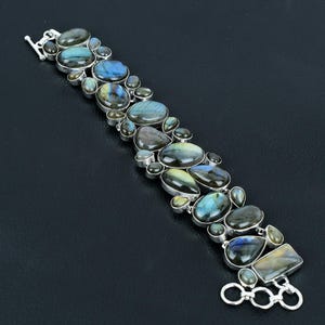 Puede incluir: Una pulsera de plata con una fila de piedras preciosas de labradorita iridiscente en varias formas y tamaños. La pulsera tiene un cierre de langosta y un extensor de cadena.