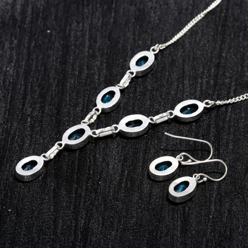 May include: A silver necklace with a pendant featuring a cluster of blue gemstones. The necklace is paired with matching earrings with a single blue gemstone.