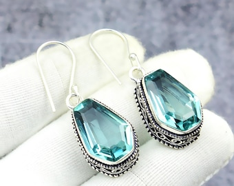 May include: A pair of silver earrings featuring large, rectangular, light blue gemstones. The gemstones are set in ornate, dark-toned metal frames with decorative edging. The earrings have a simple hook design.