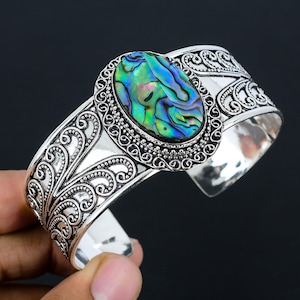Puede incluir: Pulsera de brazalete de plata con una incrustación de concha de abulón iridiscente. La pulsera tiene un diseño floral detallado.