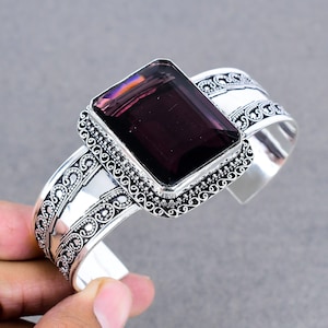 May include: A silver cuff bracelet featuring a large, rectangular, deep purple gemstone. The gemstone is set in a decorative frame with intricate detailing. The bracelet has a wide band with ornate scrollwork patterns.