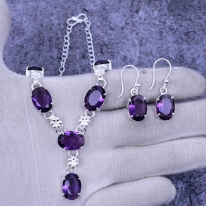 May include: A silver necklace and earrings set with oval-shaped purple gemstones. The necklace has a Y-shaped design with a smaller gemstone pendant at the bottom.