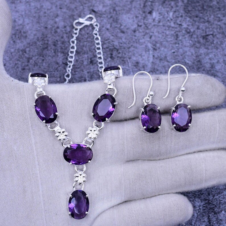 May include: A silver necklace and earrings set with oval-shaped purple gemstones. The necklace has a Y-shaped design with a smaller gemstone pendant at the bottom.