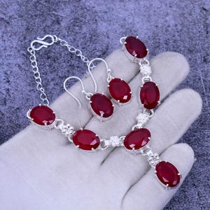May include: A silver necklace and earrings set with red oval-shaped gemstones. The necklace has a pendant with a larger gemstone and a chain with smaller gemstones.