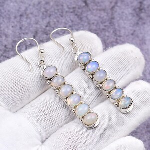 May include: A pair of silver earrings featuring a row of oval-shaped opals. The opals have a milky white base with flashes of iridescent color. The earrings are displayed on a white surface.
