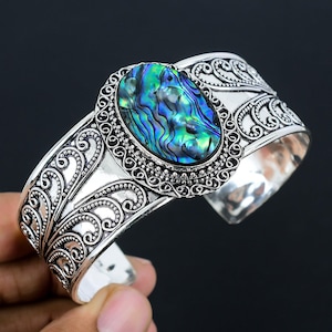 Könnte beinhalten: Silbernes Armreif-Armband mit einem ovalen Einlegeteil aus Abalone-Muschel. Das Armband hat ein dekoratives Blumenmuster.