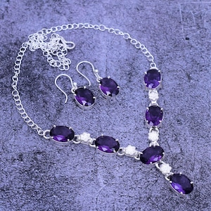 May include: A silver necklace and earrings set with purple oval-shaped gemstones. The necklace has a delicate chain and features five gemstones with small silver flower accents between each stone. The earrings are simple and feature a single purple gemstone.