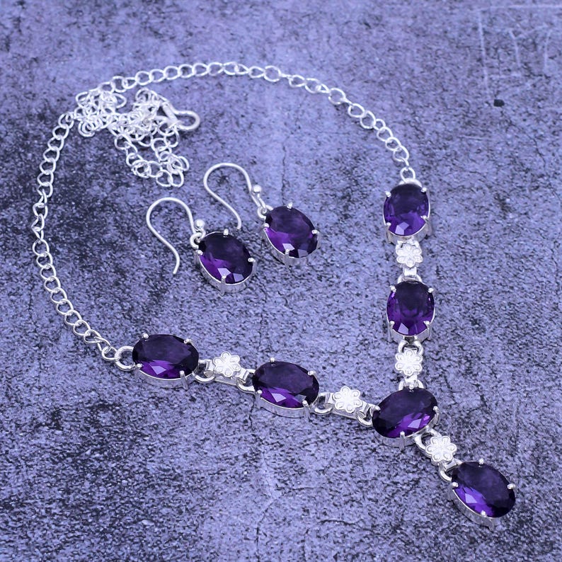 May include: A silver necklace and earrings set with purple oval-shaped gemstones. The necklace has a delicate chain and features five gemstones with small silver flower accents between each stone. The earrings are simple and feature a single purple gemstone.