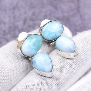 Puede incluir: Un par de pendientes de plata con piedras de larimar azul claro de forma ovalada y de lágrima. Las piedras tienen un aspecto veteado con tonos blancos y azules. Los pendientes están sobre una superficie blanca.