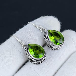 May include: A pair of silver dangle earrings featuring green pear-shaped gemstones. The earrings have an intricate filigree design around the gemstones.