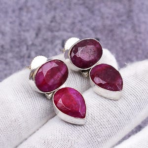 Puede incluir: Un par de pendientes de plata de ley con piedras preciosas de rubí de forma ovalada y de lágrima. Los rubíes son de color rojo intenso y están engastados en biseles de plata pulida. Los pendientes se muestran sobre una superficie blanca.