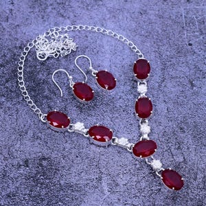 May include: A silver necklace and earrings set featuring red oval-shaped gemstones. The necklace has a delicate chain with a pendant and multiple red stones.