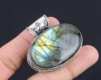 Puede incluir: Un colgante de plata con una piedra preciosa de labradorita de forma ovalada. La piedra preciosa presenta un brillo iridiscente verde, azul y dorado. El colgante tiene una argolla de plata adornada.