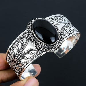 Op de afbeelding: Een zilveren armband met een sierlijk ontwerp met een zwarte ovale edelsteen. De armband heeft een brede band met een bloemmotief.