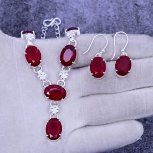 May include: A silver necklace and earrings set with red oval-shaped gemstones. The necklace has a pendant with a larger gemstone and a chain with smaller gemstones.