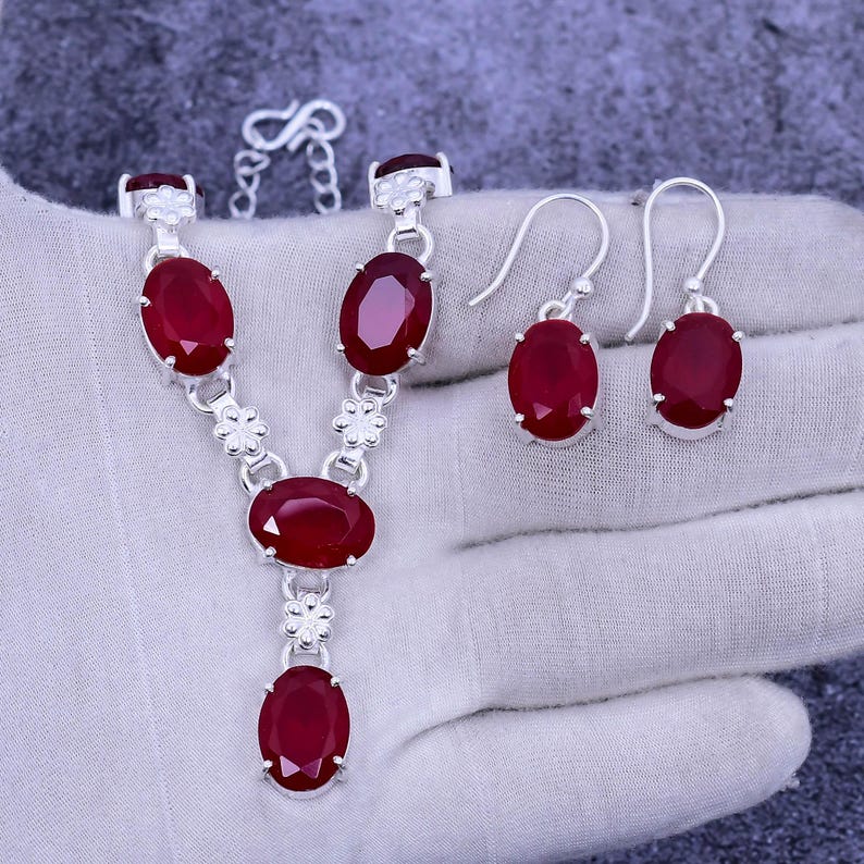 May include: A silver necklace and earrings set with red oval-shaped gemstones. The necklace has a pendant with a larger gemstone and a chain with smaller gemstones.
