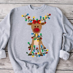 K&ouml;nnte beinhalten: Ein graues Sweatshirt mit einem Cartoon-Rentier, das eine rote Schleife tr&auml;gt und in bunten Weihnachtslichtern verheddert ist.