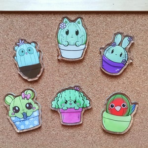 Könnte beinhalten: Sechs bunte, kaktusförmige Acryl-Anhänger mit niedlichen Gesichtern. Jeder Anhänger zeigt ein anderes Kaktusdesign mit einem Topf und einer Blume oben drauf. Die Anhänger befinden sich auf einem Korkuntergrund.