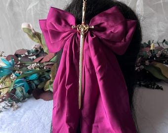 May include: A deep magenta bow hair accessory with a gold-colored sword-shaped pin. The bow is made of a silky fabric and is attached to a black hair. The sword pin has intricate detailing.