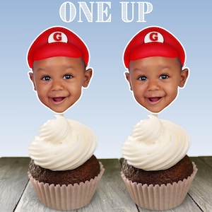 May include: Two cupcakes with white frosting and chocolate cake. Each cupcake has a red and white Mario hat with a letter G on top. The hats have a picture of a baby's face inside them. The text 