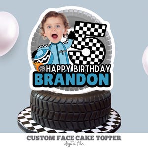 May include: A custom face cake topper featuring a black tyre and a chequered flag design. The topper includes a photo of a child in a blue racing suit with the text "Happy Birthday Brandon".
