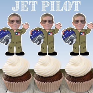 May include: Three cupcake toppers featuring a cartoon pilot wearing a blue and white helmet and a green and brown flight suit. The pilot is holding a blue and white helmet with the word "Maverick" on it. The toppers are placed on top of chocolate cupcakes with white frosting.
