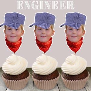 May include: Three cupcakes decorated with engineer-themed toppers. Each topper features a child wearing a blue and white striped engineer hat and a red bandana. The cupcakes are frosted with white frosting.
