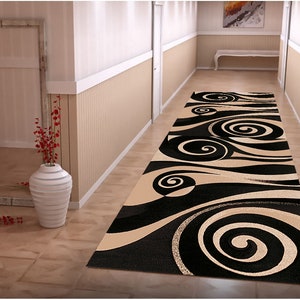 May include: A black and beige patterned runner rug with a swirling design. The rug is laid out in a hallway with white walls and a wooden floor.