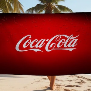 May include: A red Coca-Cola beach towel with the Coca-Cola logo in white, hanging on a washing line. The towel is displayed on a sandy beach with palm trees and the sea in the background.