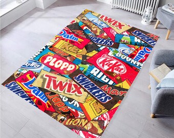 May include: A rectangular area rug featuring a collage of colorful candy bar wrappers, including Snickers, Twix, and Kit Kat. The rug has a mix of red, blue, yellow, and brown hues, creating a vibrant and playful design. The rug is placed on a light gray floor.