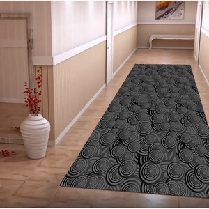 May include: A black and white patterned runner rug with a circular design. The rug is laid out in a hallway with white walls and a wooden floor.