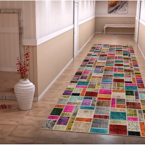 Op de afbeelding: Een kleurrijk rechthoekig tapijt met een patchwork-ontwerp, met verschillende rechthoekige vormen in roze, blauw, groen, paars en oranje. Het tapijt ligt in een gang met lichtgekleurde muren en een houten vloer.