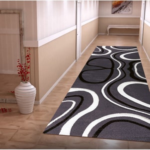 May include: A gray and white runner rug with a modern abstract design of black and white wavy lines. The rug is laid out in a hallway with white walls and a wooden floor.