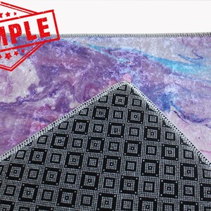 May include: A close-up of a purple and pink abstract rug with a black and white geometric pattern on the back. The rug has a gray border and is made of a soft, plush material.