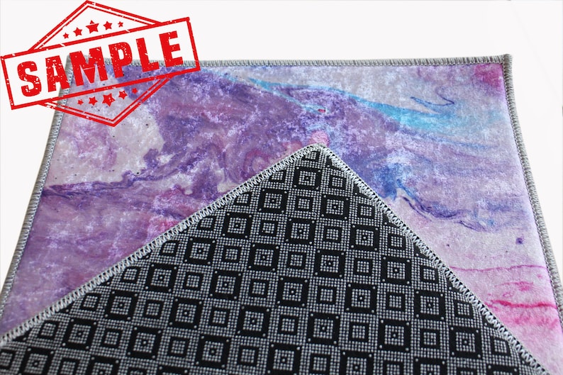 May include: A close-up of a purple and pink abstract rug with a black and white geometric pattern on the back. The rug has a gray border and is made of a soft, plush material.