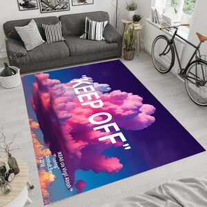 Può includere: Un tappeto viola e rosa con un design a nuvole bianche e il testo "KEEP OFF" in bianco. Il testo "IKEA® c/o Virgil Abloh™" è incluso anche sul tappeto, insieme al testo "Flooring" e "Almhult, Svezia c.2019".