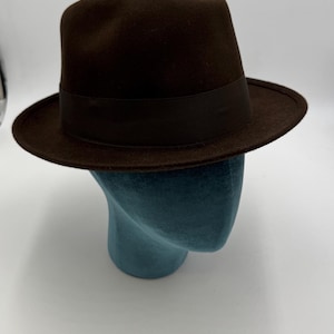 Op de afbeelding: Een bruine fedora hoed met een donkerbruine band. De hoed wordt getoond op een turquoise mannequinhoofd. De hoed heeft een klassiek ontwerp met een licht ingedeukte kroon en een rand die naar beneden buigt. De hoed is gemaakt van een viltachtig materiaal.