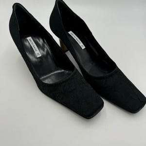 May include: A pair of black square-toe pumps with textured fabric. The heels are a light brown color. The brand name "BRUNO MAGLI" is printed on a white label inside each shoe.
