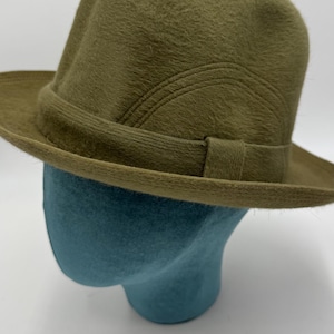 May include: Olive green fedora hat with a structured crown and a band around the base. The hat is displayed on a blue head form against a white background. The hat's brim is slightly curved.