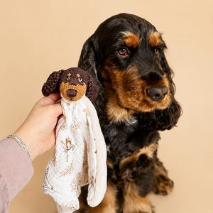 Puede incluir: Una manta de seguridad de ganchillo con una cabeza de perro marrón y tostada con orejas caídas unida a un cuadrado de tela de muselina blanca. La tela tiene un delicado estampado botánico marrón y un reverso de material suave y texturizado.