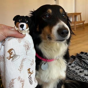May include: A white muslin security blanket featuring a repeating brown botanical and mushroom print, topped with a crocheted black, brown, and white dog head with a cream-colored ruffled collar, positioned next to a black, white, and brown dog.