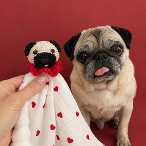 Può includere: Un giocattolo all'uncinetto a forma di testa di carlino attaccato a una coperta di tessuto bianco con un motivo di piccoli cuori rossi. La testa del carlino ha il muso beige, le orecchie nere e il naso nero con una minuscola lingua rosa.