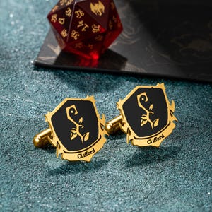 Könnte beinhalten: Ein Paar goldfarbene Manschettenknöpfe mit einem schwarzen Mittelteil, das ein stilisiertes Blumendesign zeigt. Das Wort "Clifford" ist auf der Unterseite jedes Manschettenknopfes eingraviert. Ein roter und goldener Würfel ist im Hintergrund zu sehen.