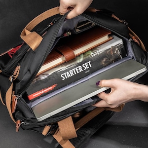 K&ouml;nnte beinhalten: Ein schwarzer Canvas-Rucksack mit braunen Lederelementen und einer Rei&szlig;verschluss&ouml;ffnung oben. Der Rucksack ist gef&uuml;llt mit B&uuml;chern, einem Laptop und einer Brettspielbox mit der Aufschrift "Starter Set" und "Dungeons & Dragons".